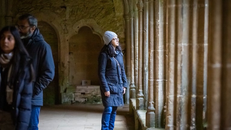 A visitor looks out of the window in the Cloister at Lacock during winter. They are wearing a warm hat and coat, and looking pensive.